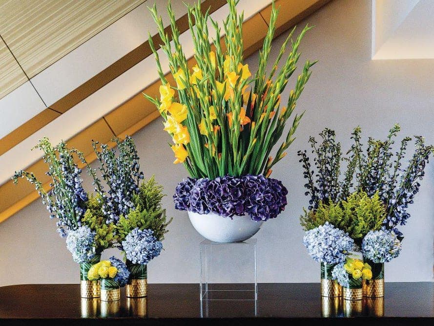 Grouped South Florals memorial arrangements in blue, yellow and purple on a dark console table.